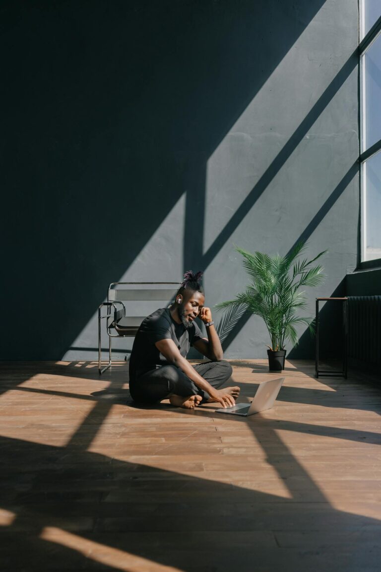 A young man sitting on a wooden floor, using a laptop in bright, modern loft space with large windows.