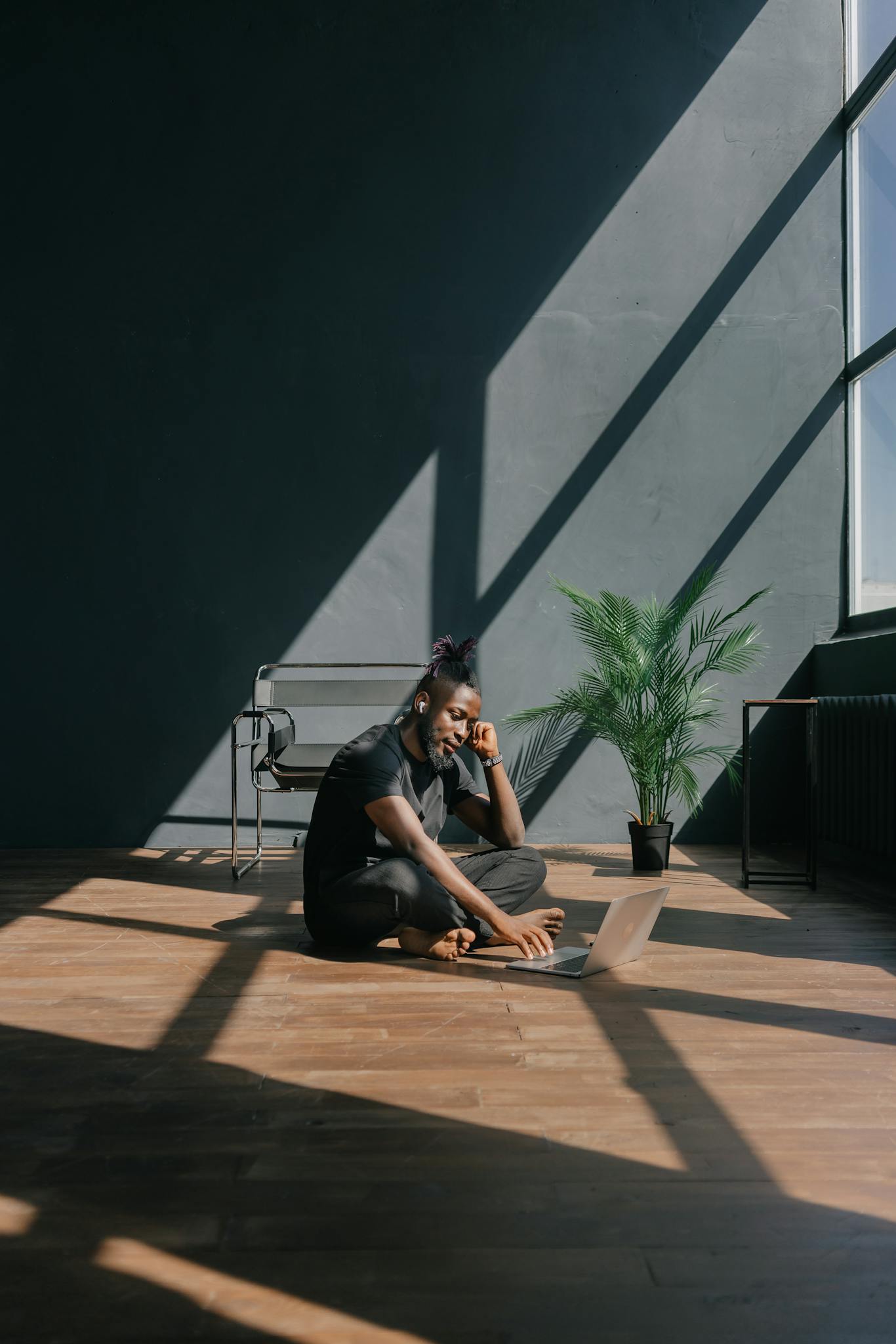 A young man sitting on a wooden floor, using a laptop in bright, modern loft space with large windows.