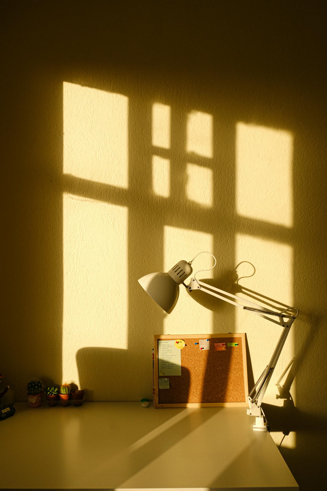 An artistic desk setup featuring a lamp casting shadows and a cork board in warm lighting.